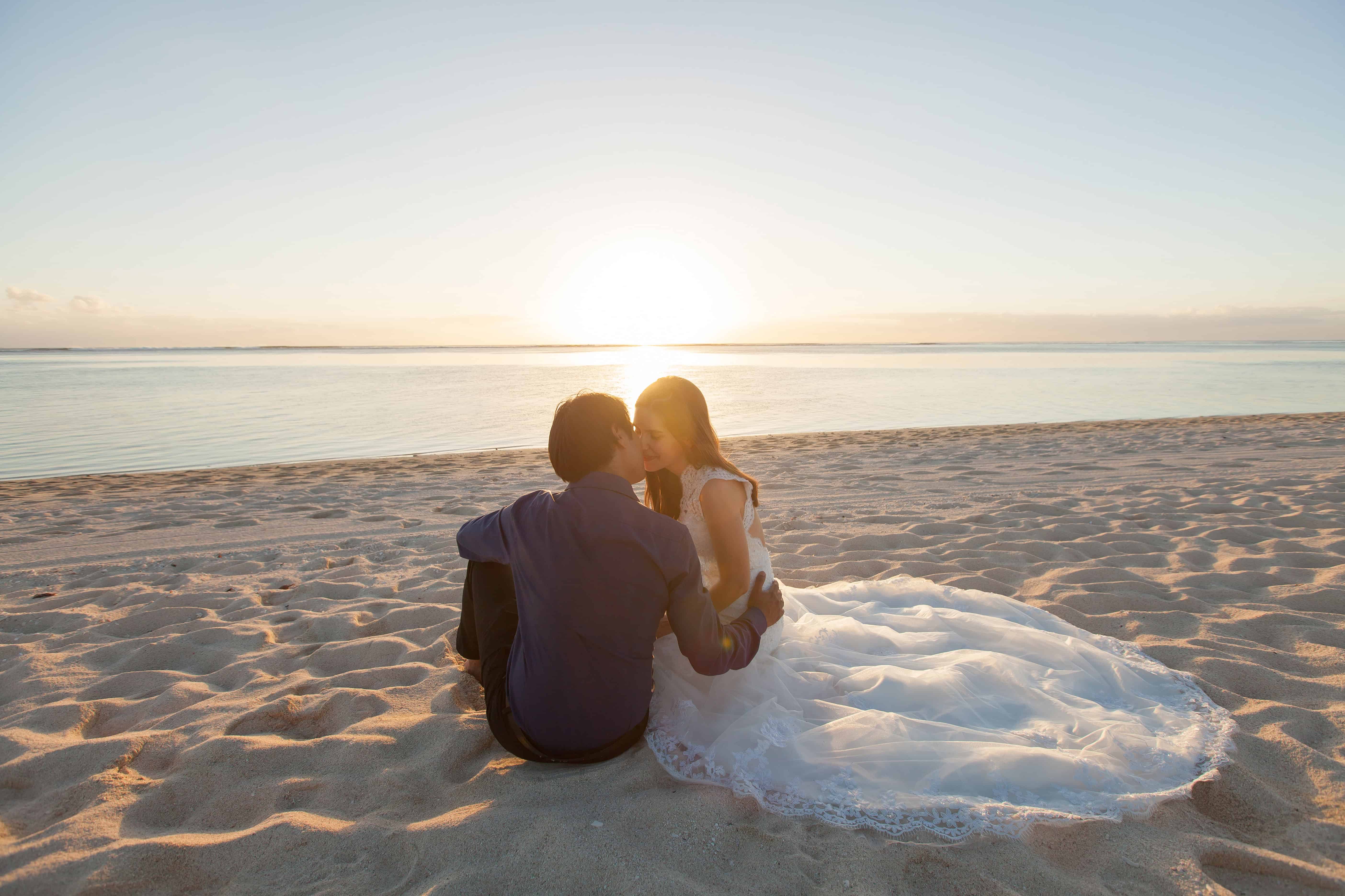 Braut und Bräutigam sitzen bei Sonnenuntergang am Strand und küssen sich, das Brautkleid liegt im Sand neben dem Paar.